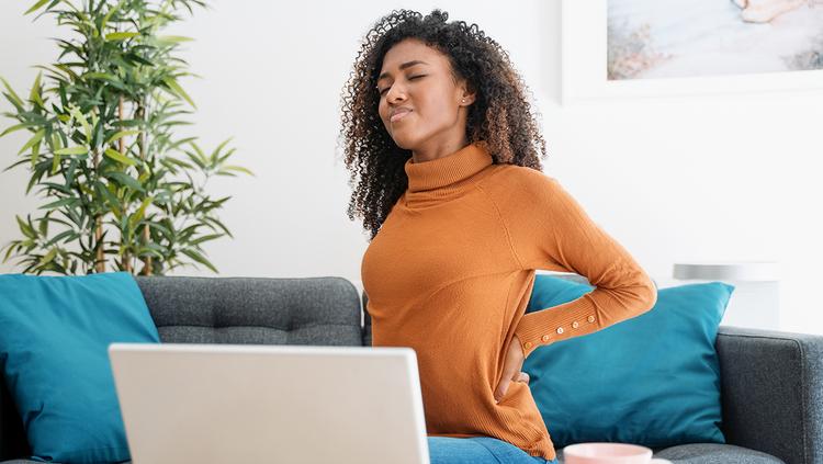 Woman seated on a sofa with a laptop, holding her lower back.